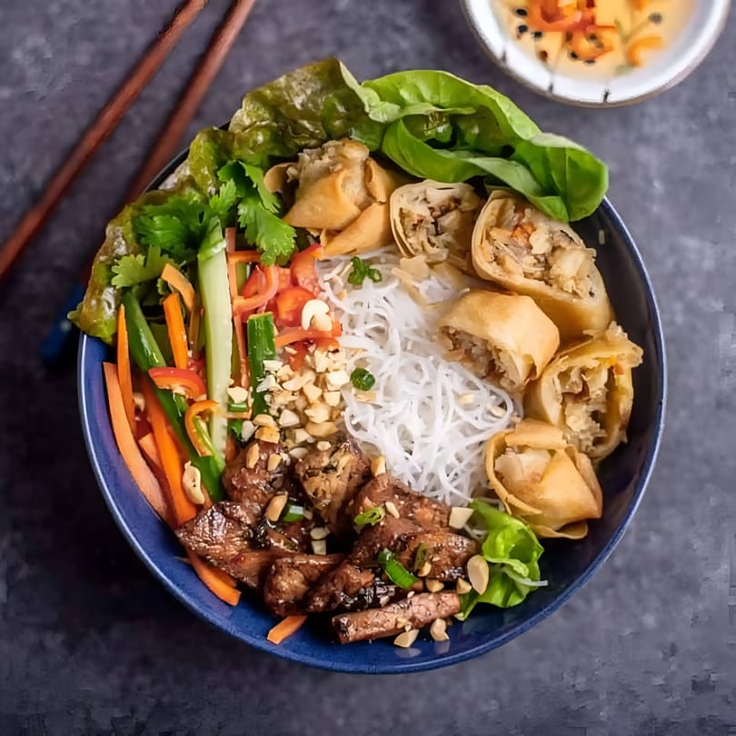 A vibrant Vietnamese vermicelli bowl with grilled pork, crispy spring rolls, fresh vegetables, and peanuts.
