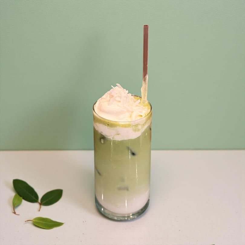 Iced matcha latte with whipped cream and shredded coconut, with green leaves on a white surface.