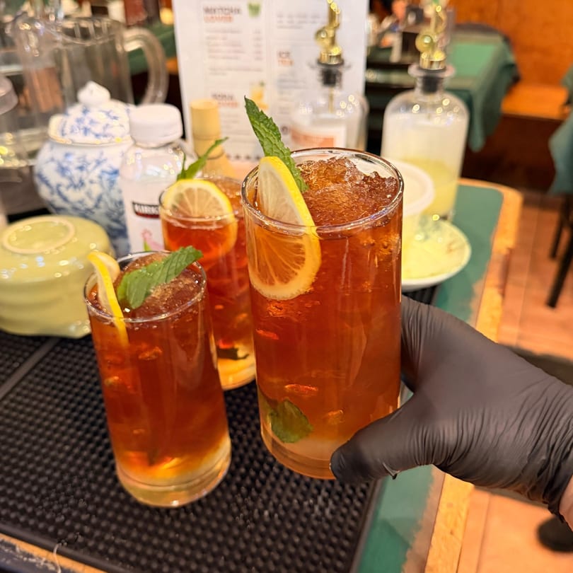 A gloved hand holds a tall glass of iced tea with lemon and mint, next to another glass.