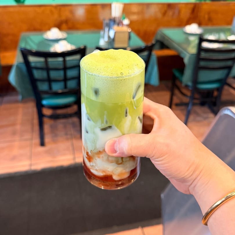 Hand holding a vibrant layered drink with green foam, white liquid, and red syrup in a restaurant.