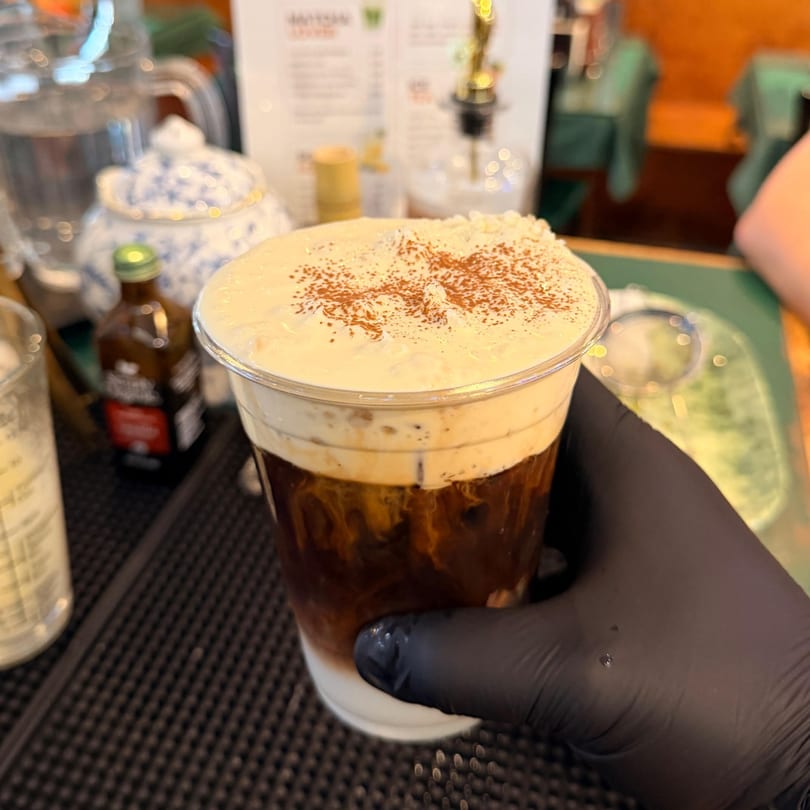 A hand in a black glove holds a Vietnamese iced coffee with cream and cocoa powder.