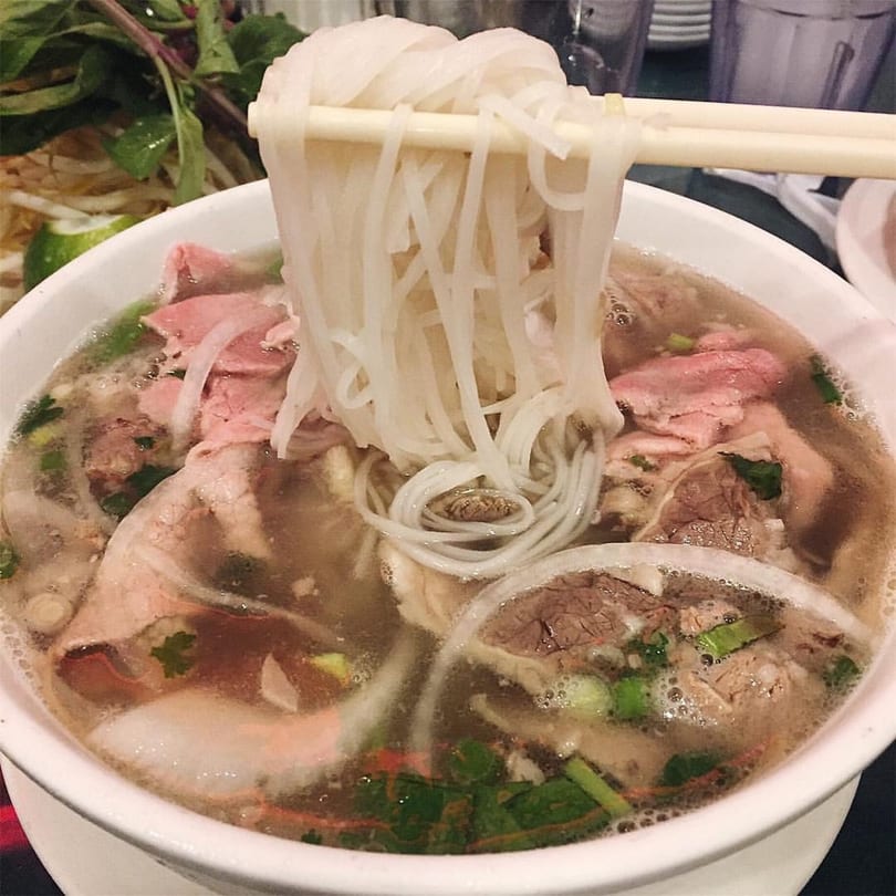 Chopsticks lift white rice noodles from a bowl of pho with beef, broth, and herbs.