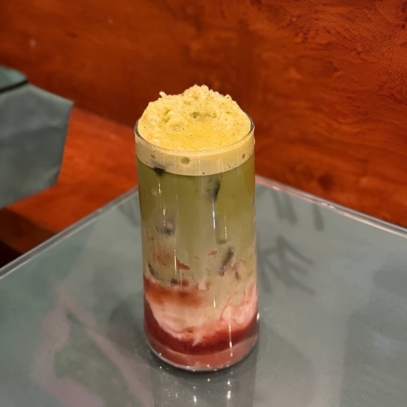 Tall glass of layered green matcha and pink strawberry drink with ice and foam on a table.