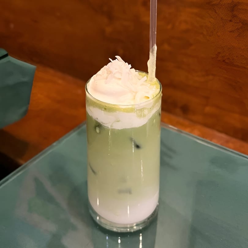 Iced green drink with white cream and coconut flakes on top, with a straw.