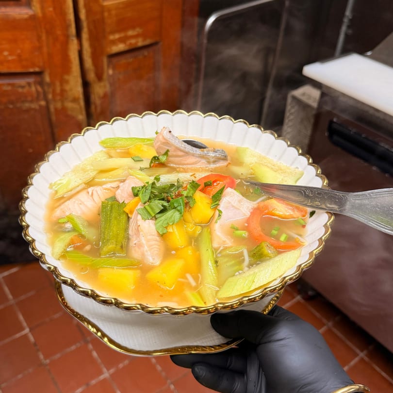 A steaming bowl of Vietnamese salmon soup with vegetables, held by a gloved hand.