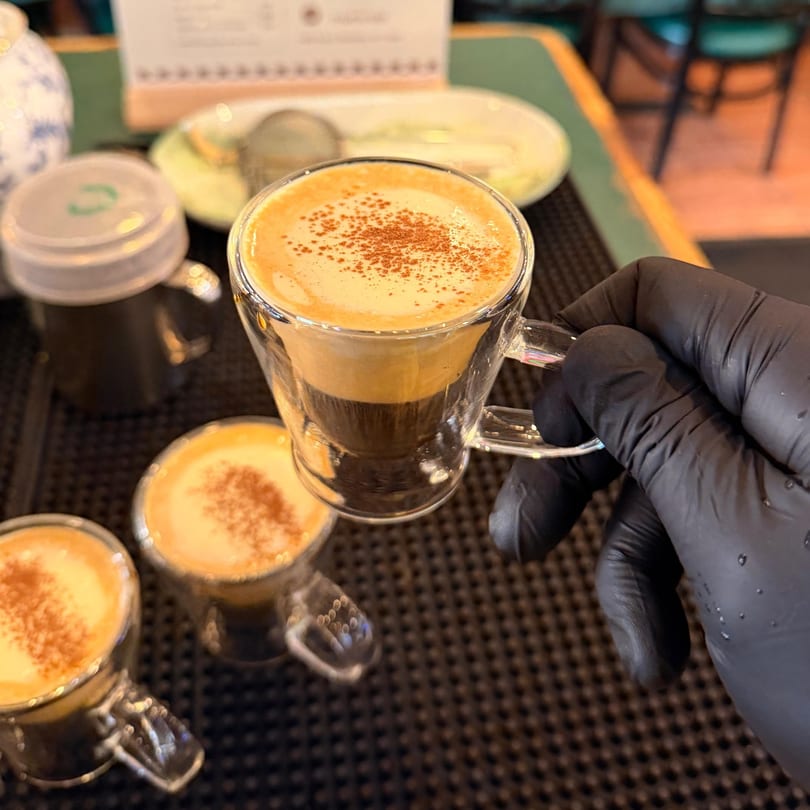 A gloved hand holds a small glass cup of Vietnamese egg coffee with cinnamon sprinkles.
