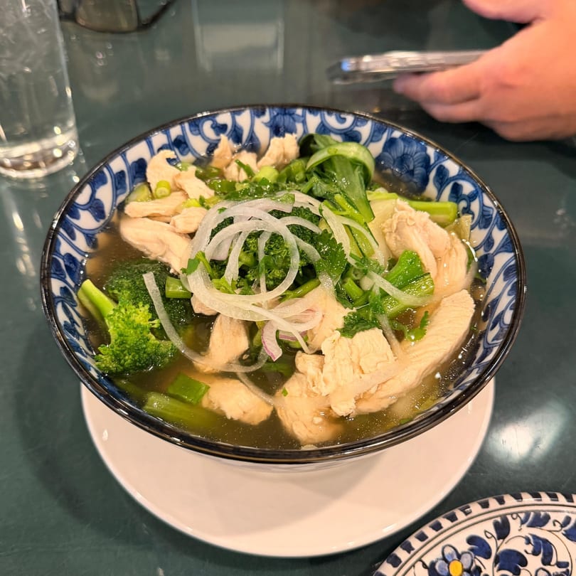 Pho Hoai chicken pho with broccoli, green onions, and sliced onions in a blue and white bowl.